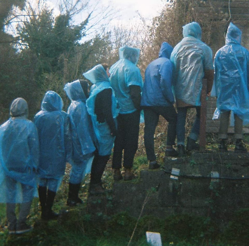 grainy, murky, film photograph. members of Trá Pháidín face away from the camera, on a moss-covered staircase in the middle of a thick forest. all members of the band are wearing translucent blue rain ponchos with the hoods up.